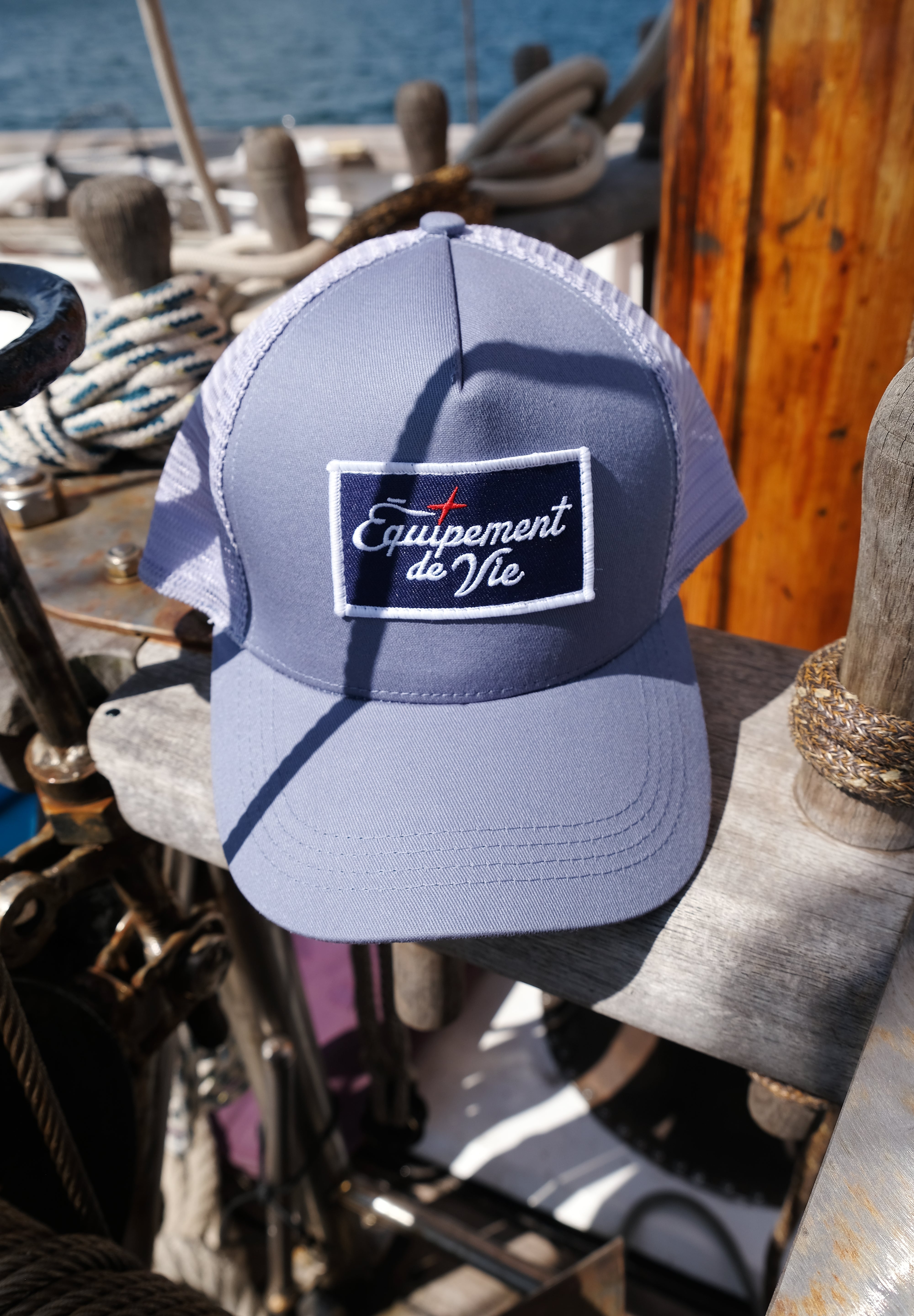 Trucker Cap in Dust Blue and Grey with Équipement De Vie patch, on deck of a classic yacht