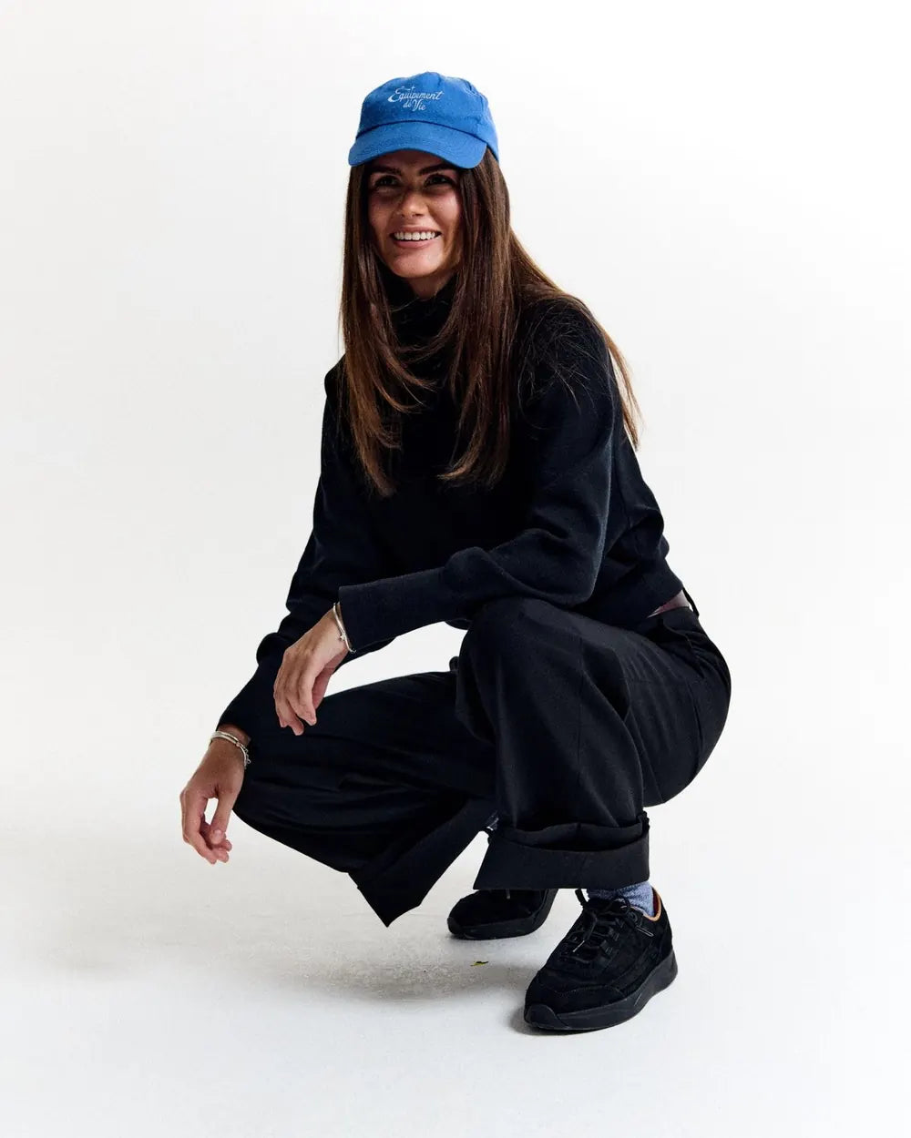 Woman crouching in a black outfit and blue Équipement De Vie cap