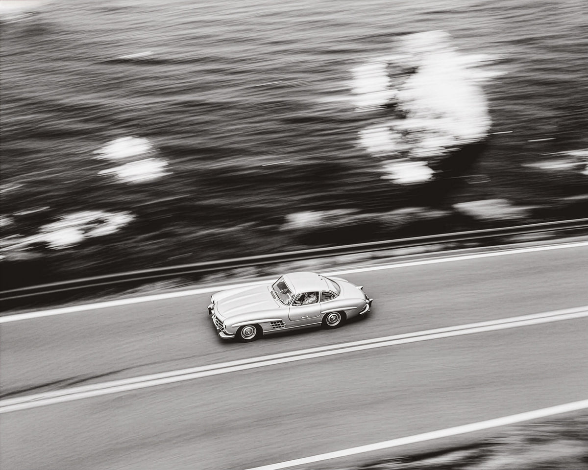 Classic silver sports car speeding along a coastal road, black and white
